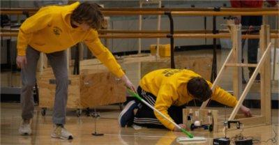 two students in yellow work on floor during robotics event, one holding swifter mop